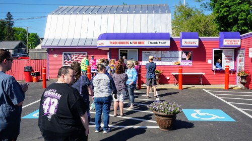A line of people waiting to order at a colorful fast-food restaurant with a red exterior and a silver roof.