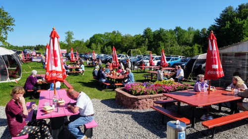 Outdoor dining area with people at picnic tables, red umbrellas, and a green landscape in the background.