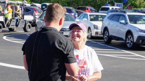 A smiling woman in a cap talks to a man in a black shirt at a busy outdoor event with cars in the background.