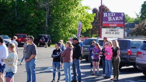 A crowd of people stands in a parking lot near a sign for Red Barn, surrounded by trees and parked vehicles.