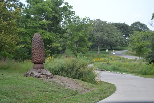 A large, pinecone-shaped sculpture stands beside a winding path in a green, wooded area.