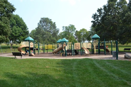 A playground with slides and climbing structures, surrounded by green grass and trees on a sunny day.