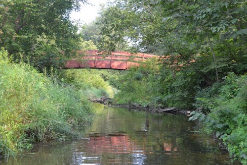 A rusted bridge spans a serene creek, surrounded by lush greenery and trees reflecting in the calm water.