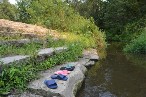A pair of colorful sandals rests on a stone by a calm stream, surrounded by lush greenery.