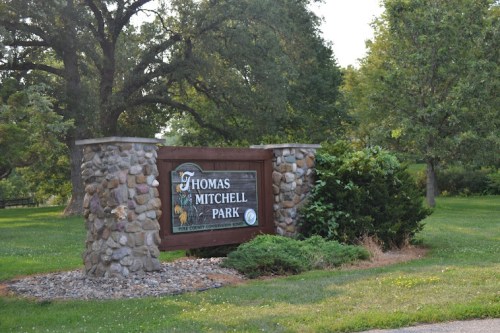 Sign for Thomas Mitchell Park, surrounded by greenery and trees, with a stone base and a grassy area in the background.