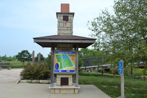 A park information kiosk with a map, surrounded by greenery and a bridge in the background. Accessible sign nearby.