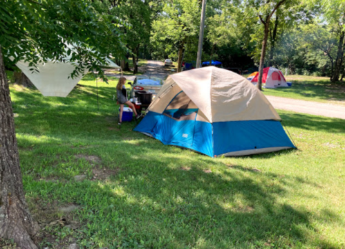 A blue and tan tent set up in a grassy campsite with trees, and a person sitting nearby at a camping table.