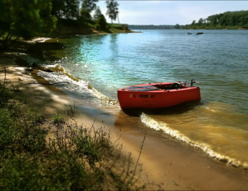 A bright orange boat rests on a sandy shore by a calm lake, surrounded by trees and gentle waves.