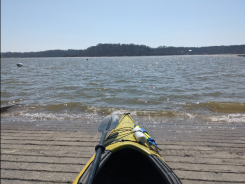A yellow kayak on a dock, facing a calm lake with gentle waves and distant trees under a clear blue sky.