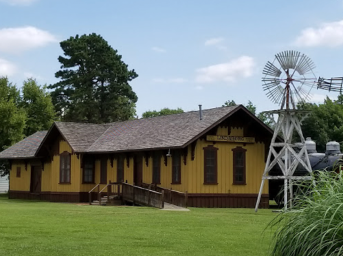 Historic yellow train station with a wooden ramp, surrounded by green grass and a windmill nearby.
