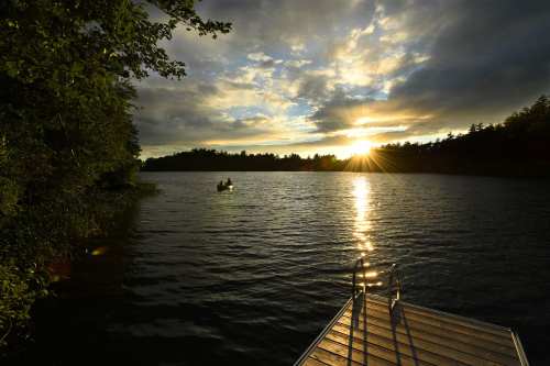 A serene lake at sunset, with a dock in the foreground and two people in a boat on the water.