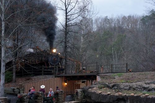 A steam train emits smoke near a wooded area, with people gathered on a deck decorated with lights.