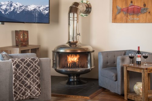 A cozy living room with a modern fireplace, two gray chairs, a wooden table with wine, and a mountain view on the TV.