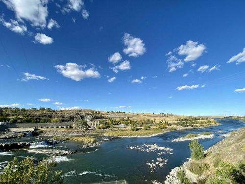 A scenic river flows through a landscape of hills and trees under a blue sky with scattered clouds.