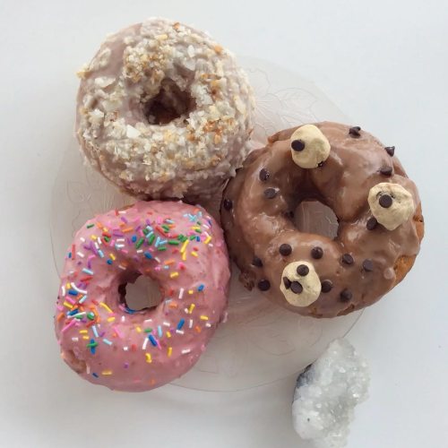Three colorful donuts on a plate: one pink with sprinkles, one chocolate with eyes, and one coconut-covered.