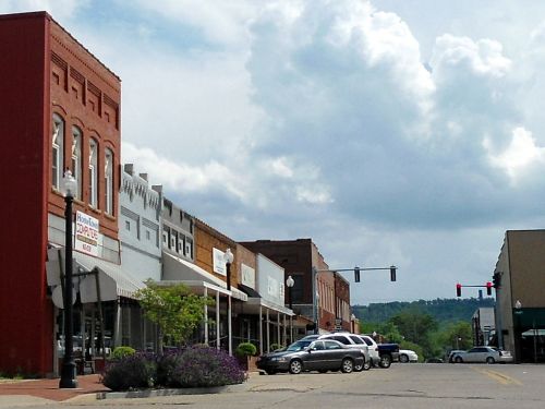 A small town street lined with brick buildings, shops, and parked cars under a cloudy sky.