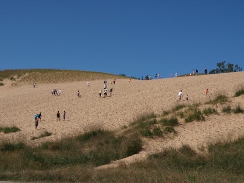 People climbing a sandy dune under a clear blue sky, with green grass in the foreground.
