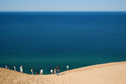 A sandy beach with people walking along the shore, overlooking a calm blue ocean under a clear sky.