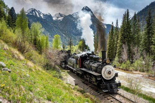 A vintage steam train chugs along a scenic mountain landscape, surrounded by trees and a river under a cloudy sky.