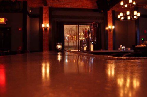 A dimly lit bar with a polished countertop, brick walls, and soft lighting creating a cozy atmosphere.