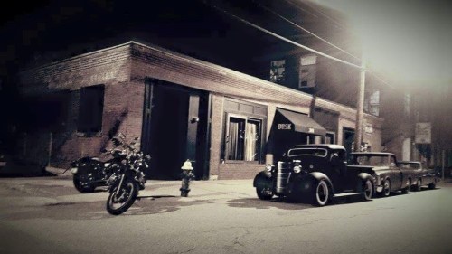 A vintage car and motorcycle parked on a dimly lit street near an old brick building at night.