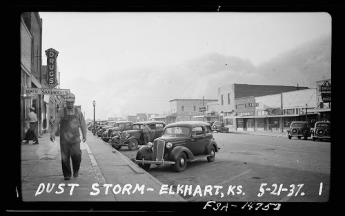A man walks along a dusty street lined with vintage cars and storefronts in Elkhart, Kansas, during a dust storm.