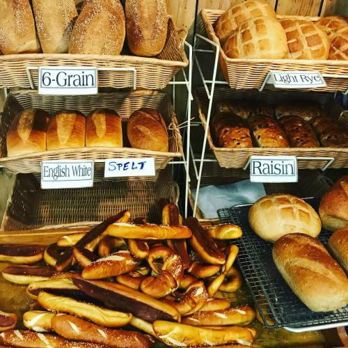 A display of various breads, including 6-grain, English white, spelt, light rye, and raisin, with pretzel rolls in front.