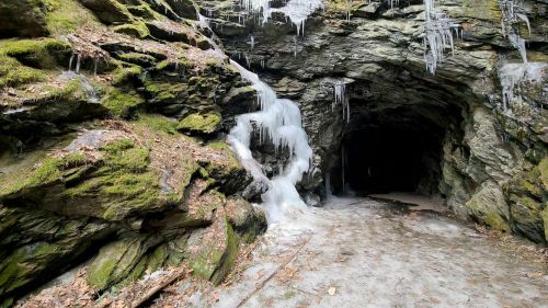 A rocky cave entrance with icicles and a small waterfall, surrounded by moss and fallen leaves.
