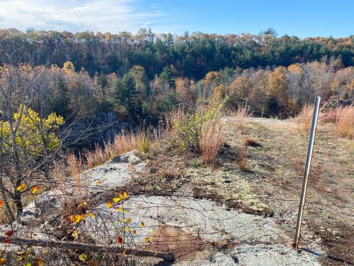 A rocky outcrop with sparse vegetation overlooks a wooded valley in autumn, featuring colorful foliage and a clear sky.