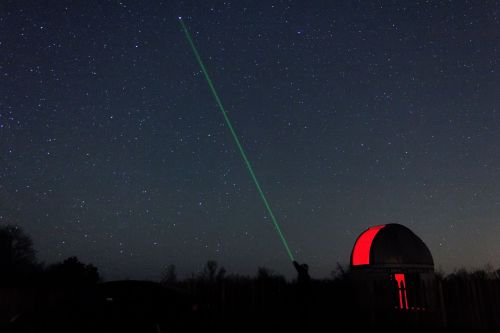A person points a green laser at the night sky from an observatory, surrounded by stars and a red-lit dome.