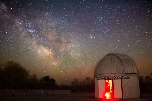 A dome-shaped observatory under a starry sky, with the Milky Way visible and a red light glowing at the entrance.