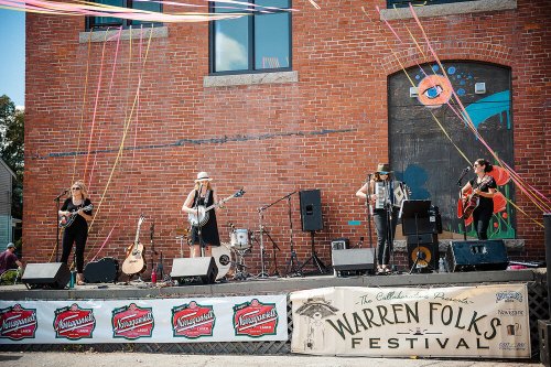 A band performs on stage at the Warren Folks Festival, with colorful decorations and instruments visible.