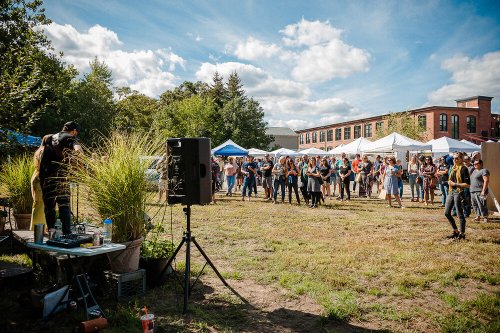 A lively outdoor event with a crowd gathered near tents, enjoying music and activities under a sunny sky.