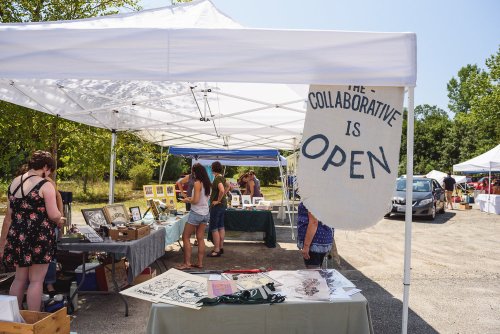 A sunny outdoor market scene with tents, people browsing art, and a sign reading "The Collaborative is Open."