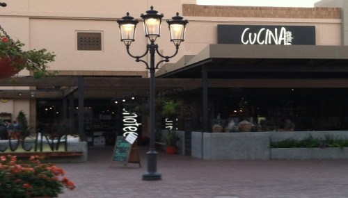 A restaurant named "Cucina" with outdoor seating, surrounded by plants and a decorative street lamp.