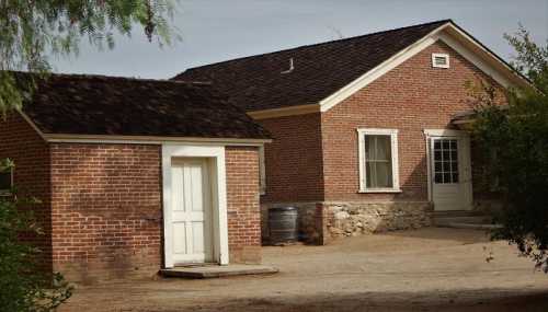 A brick house with a white door and a smaller adjacent building, set in a rural area with a dirt path.