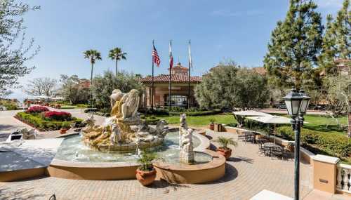 A landscaped courtyard featuring a fountain, palm trees, and a building entrance with flags in the background.