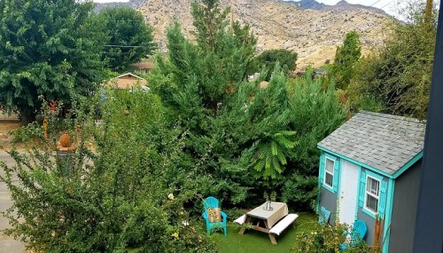 A cozy outdoor space with a small blue shed, green trees, and a table surrounded by chairs, set against a mountain backdrop.