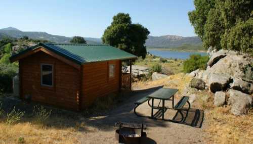 A cozy wooden cabin near a lake, surrounded by rocks and trees, with a picnic table and fire pit in the foreground.