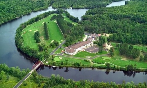 Aerial view of a golf course and resort by a river, surrounded by lush green trees and rolling hills.
