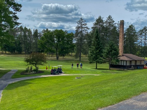 A scenic golf course with a group of golfers, a cart, and a rustic building surrounded by trees and a cloudy sky.