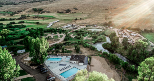 Aerial view of a campground with pools, RVs, and a river surrounded by greenery and hills. Sunlight beams in the background.
