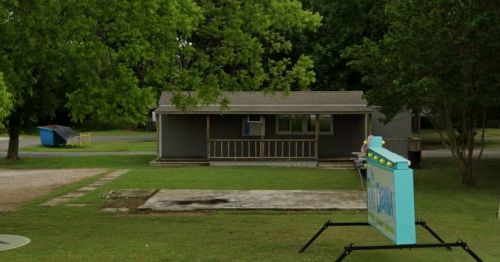 A gray house with a porch, surrounded by trees, and a sign in front on a grassy area. A dumpster is visible nearby.