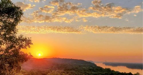 A vibrant sunset over a river, with clouds scattered in a colorful sky and lush greenery in the foreground.