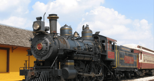 A vintage steam locomotive parked beside a yellow train station under a blue sky with fluffy clouds.