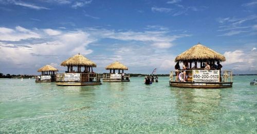 Four thatched-roof huts on stilts in clear blue water under a bright sky, with boats nearby.