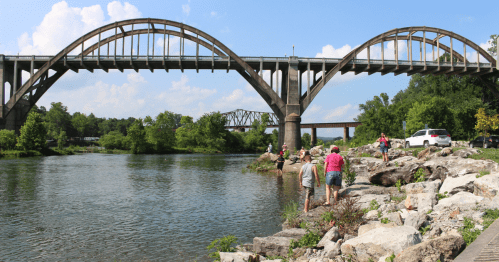 A scenic view of a river with people exploring rocky banks beneath a large arched bridge on a sunny day.