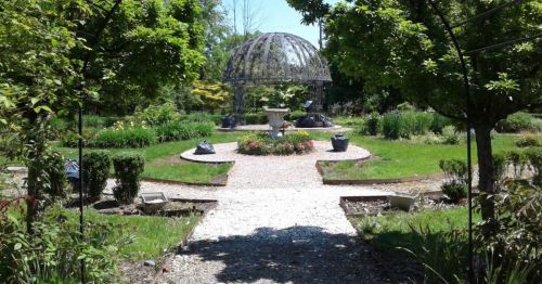 A serene garden with a central gazebo, surrounded by lush greenery and flower beds along a gravel path.