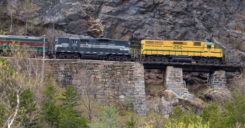 Two trains on a stone bridge, surrounded by rocky terrain and greenery. One train is black, the other yellow.