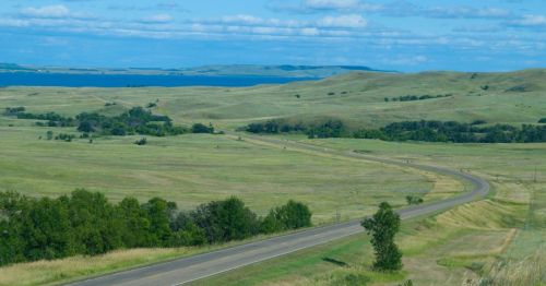A winding road through green hills, with a lake visible in the distance under a blue sky with scattered clouds.